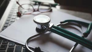 A green stethoscope rests on a laptop keyboard, with a pair of glasses and a pen blurred in the background.