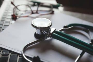 A green stethoscope rests on a laptop keyboard, with a pair of glasses and a pen blurred in the background.
