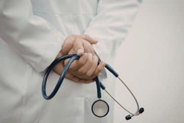 A person wearing a white lab coat holds a stethoscope behind their back.