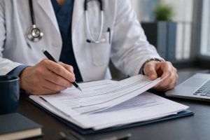 Doctor reviewing paperwork at a desk, holding a pen over documents with a laptop nearby, focusing on professional liability or insurance review